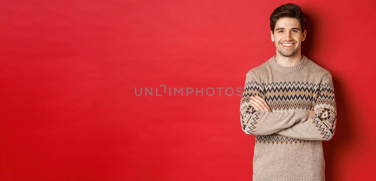 Image of handsome happy guy in christmas sweater, smiling and looking at camera, celebrating xmas holidays, standing over red background by Benzoix