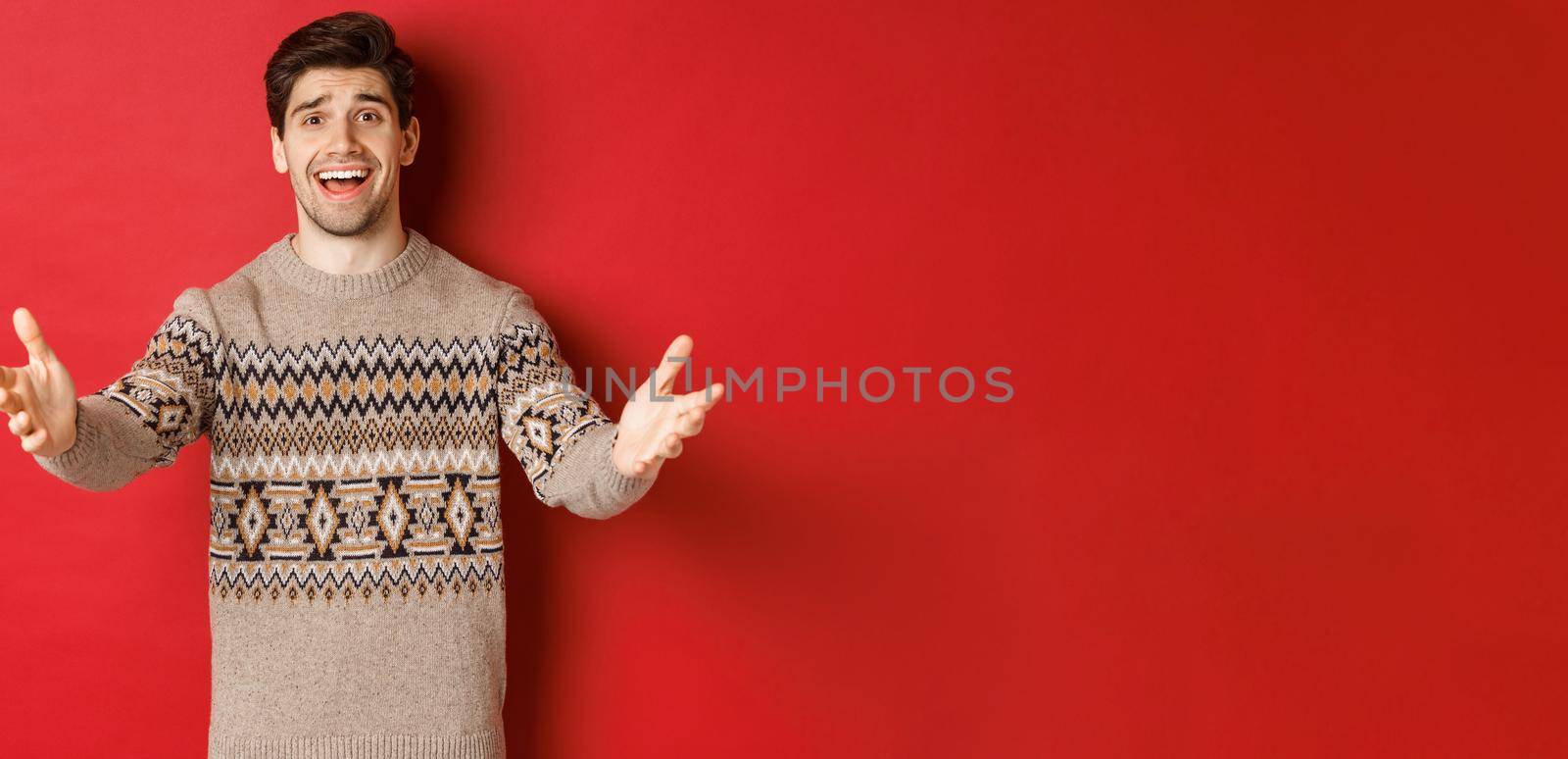 Image of happy and flattered handsome guy in christmas sweater, reaching hands to welcome guests on new year party, inviting to celebration, standing over red background by Benzoix