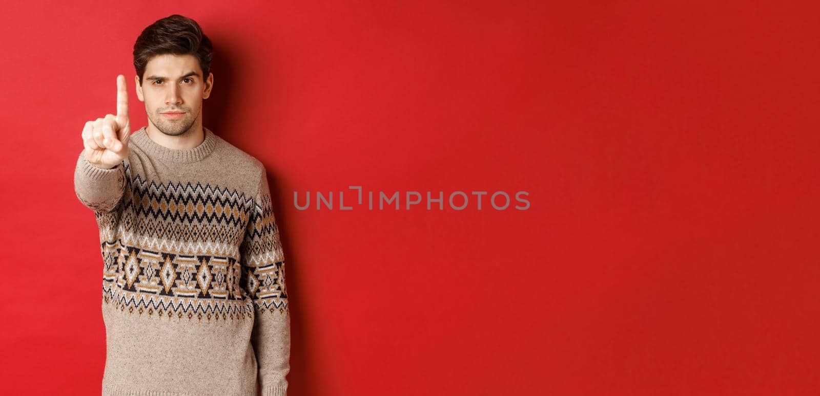 Image of serious handsome man extending one finger to stop you, telling no, give warning, standing over red background in winter sweater by Benzoix