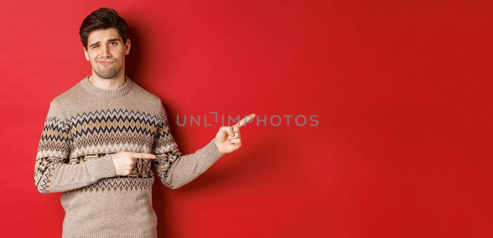 Concept of christmas celebration, winter holidays and lifestyle. Image of skeptical and displeased handsome man in xmas sweater, pointing fingers right and do not recommend product.