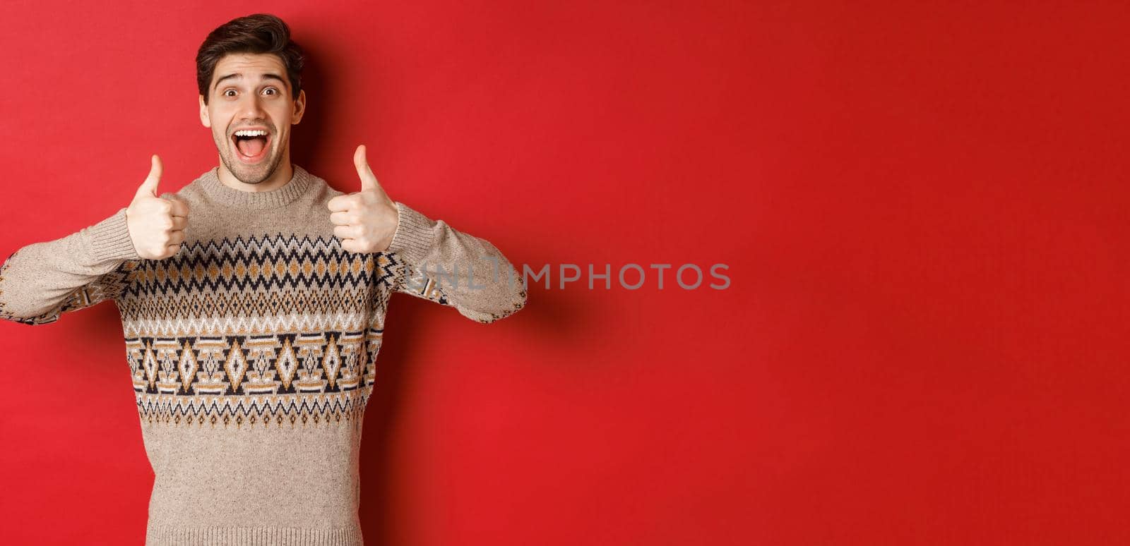 Portrait of cheerful man in christmas sweater like something good, showing thumbs-up and smiling amazed, praise new year party, standing over red background.