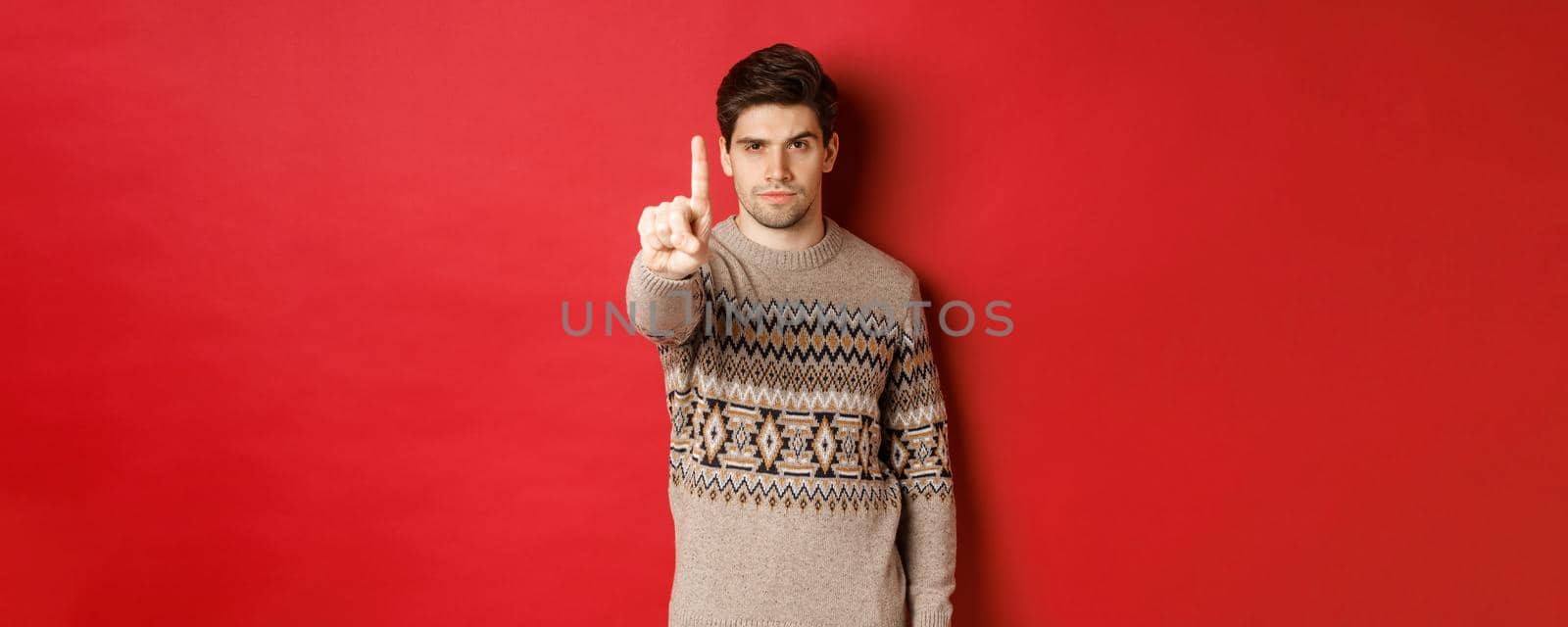 Image of serious handsome man extending one finger to stop you, telling no, give warning, standing over red background in winter sweater by Benzoix