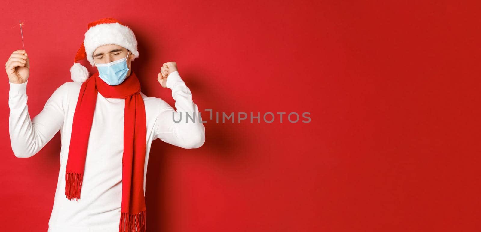 Concept of covid-19, christmas and holidays during pandemic. Happy man celebrating new year at party, wearing medical mask and santa hat, dancing with sparkler against red background by Benzoix
