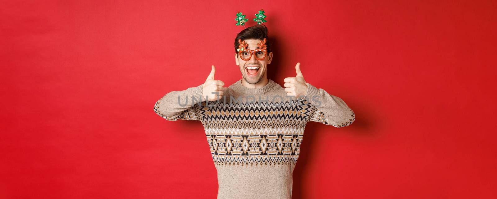 Portrait of satisfied and happy man in christmas sweater and party glasses, showing thumbs-up, wishing happy new year, standing over red background.