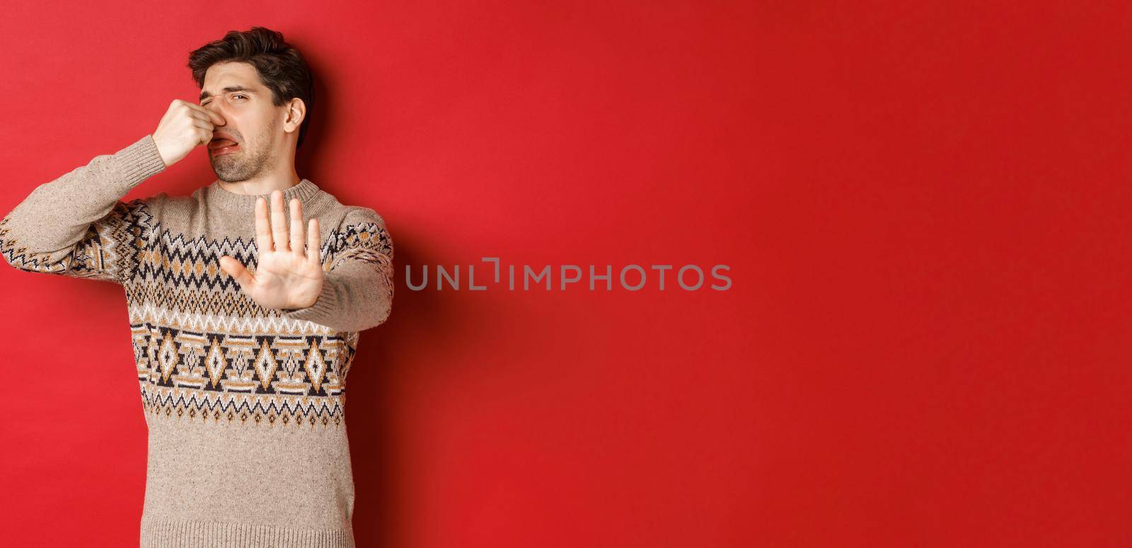 Image of disgusted handsome man in winter sweater, rejecting something with bad smell, shut nose and raising hand to decline, standing over red background by Benzoix