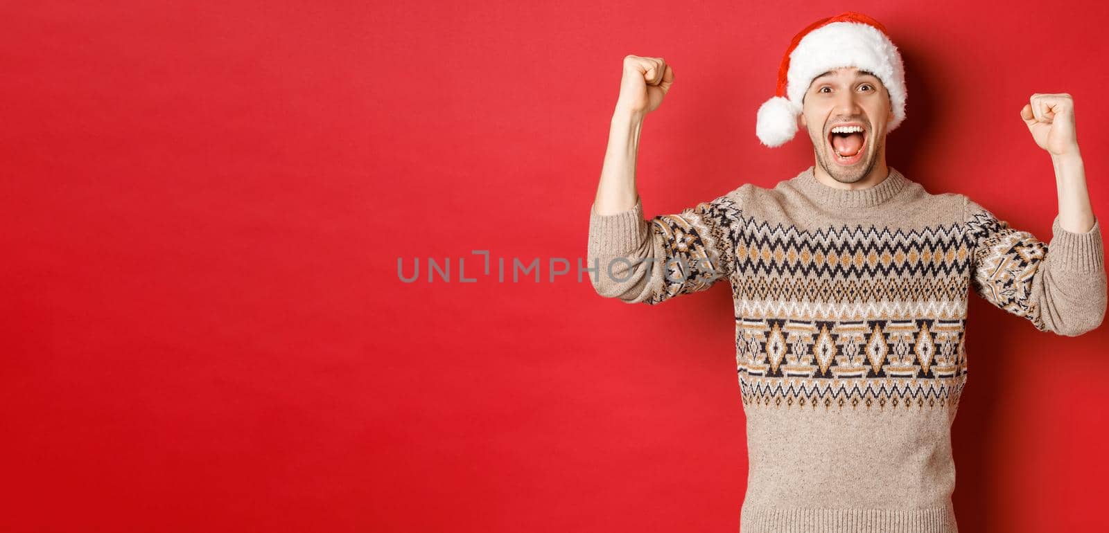Image of cheerful handsome man in swearer and santa hat, celebrating new year, triumphing or winning something, raising hands up and shouting for joy, standing over red background.