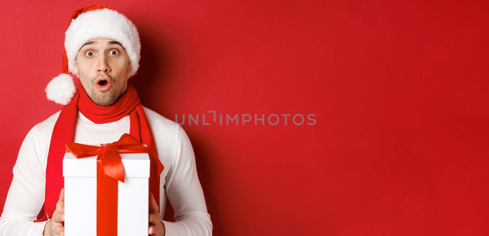 Concept of winter holidays, christmas and lifestyle. Close-up of surprised handsome guy in santa hat and scarf, looking amazed and holding new year gift, standing over red background by Benzoix