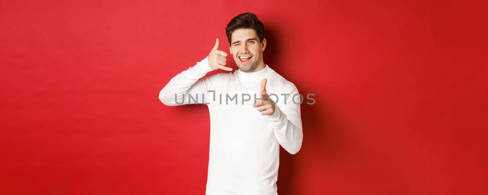 Portrait of sassy and confident handsome man, flirting with you, asking to call him, showing phone sign and pointing at camera, standing over red background by Benzoix