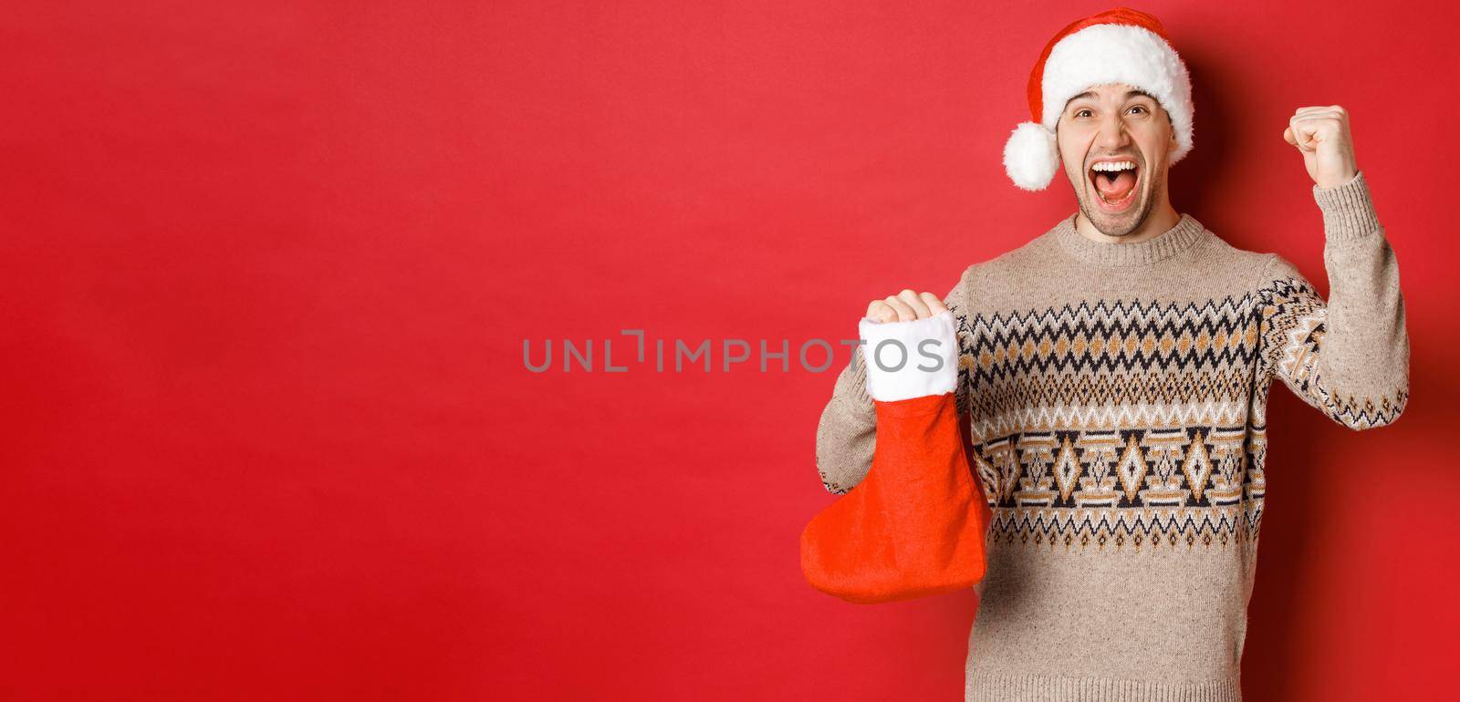 Concept of winter holidays, new year and celebration. Amazed and happy man shouting for joy, found gift inside christmas stocking and cheering, raising hand up and smiling by Benzoix