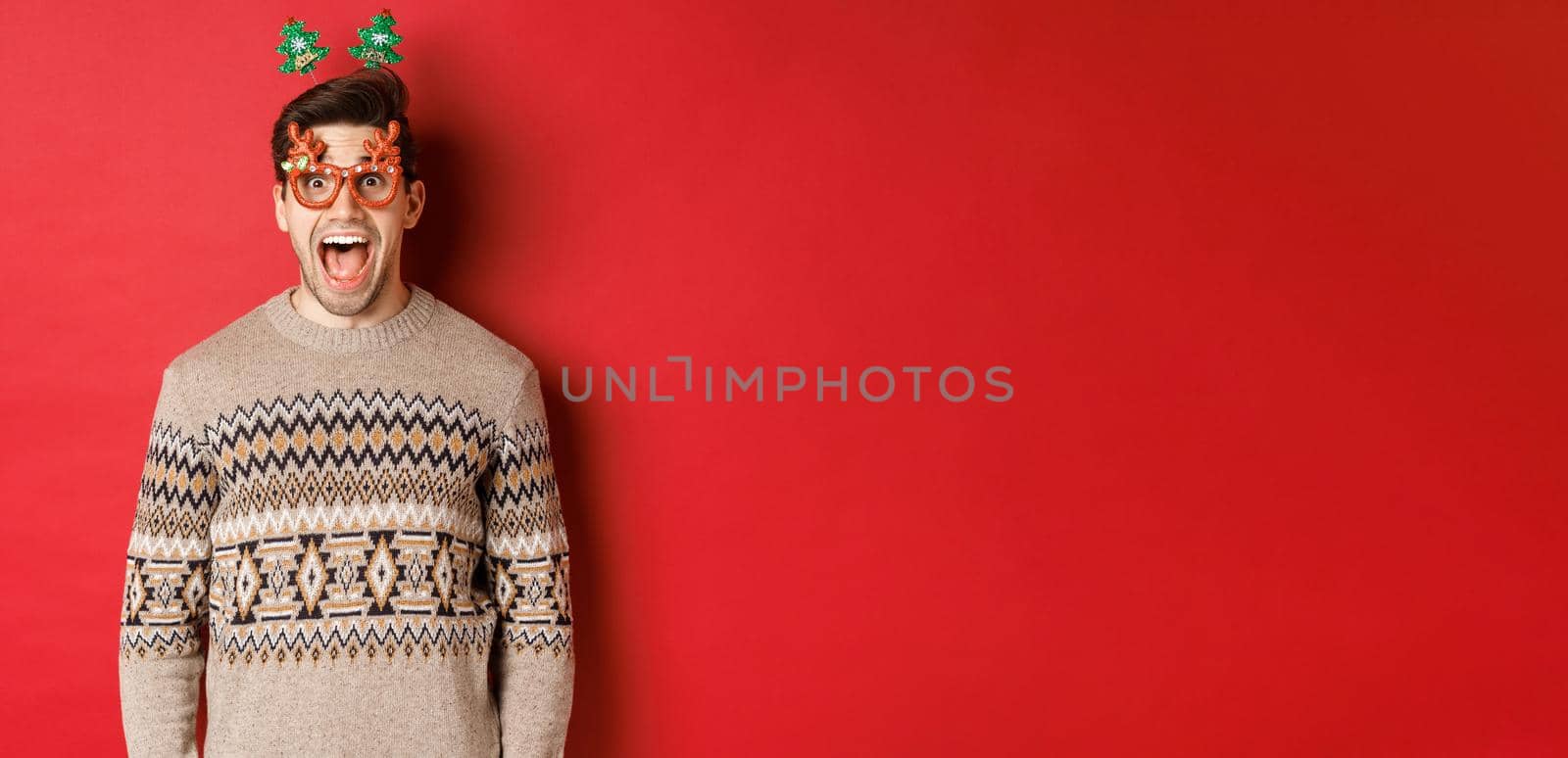 Image of surprised and excited handsome man, wearing party glasses with christmas sweater, drop jaw and looking at amazing new year promo offer, standing against red background.