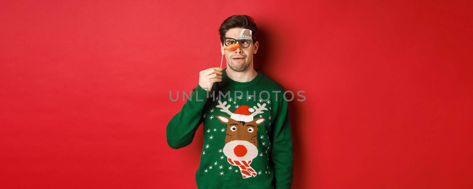 Funny man in christmas sweater and party mask, celebrating winter holidays, showing funny faces, standing over red background by Benzoix