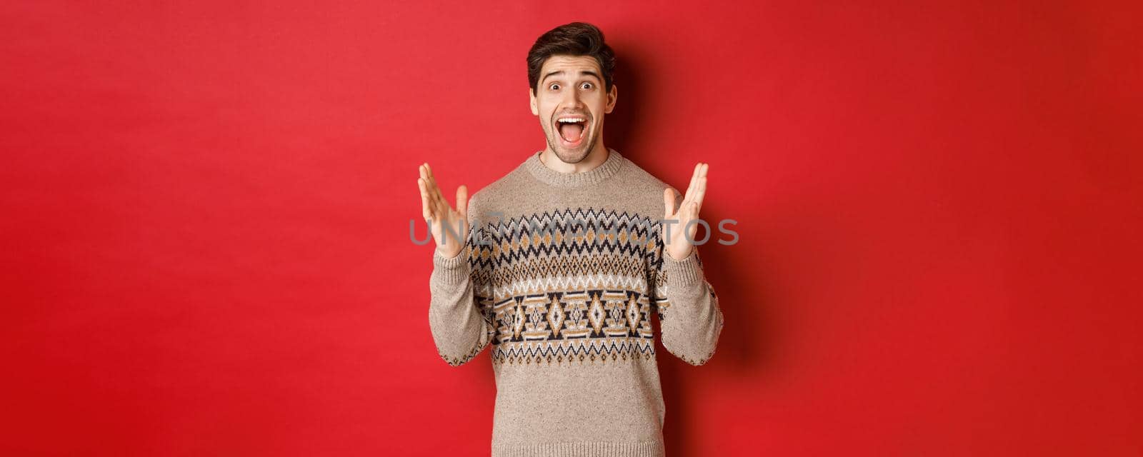 Portrait of surprised and happy handsome man, wearing christmas sweater, looking amazed, celebrating new year, standing over red background.