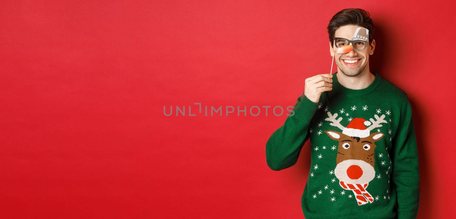 Funny man in christmas sweater and party mask, celebrating winter holidays, smiling happy, standing over red background.