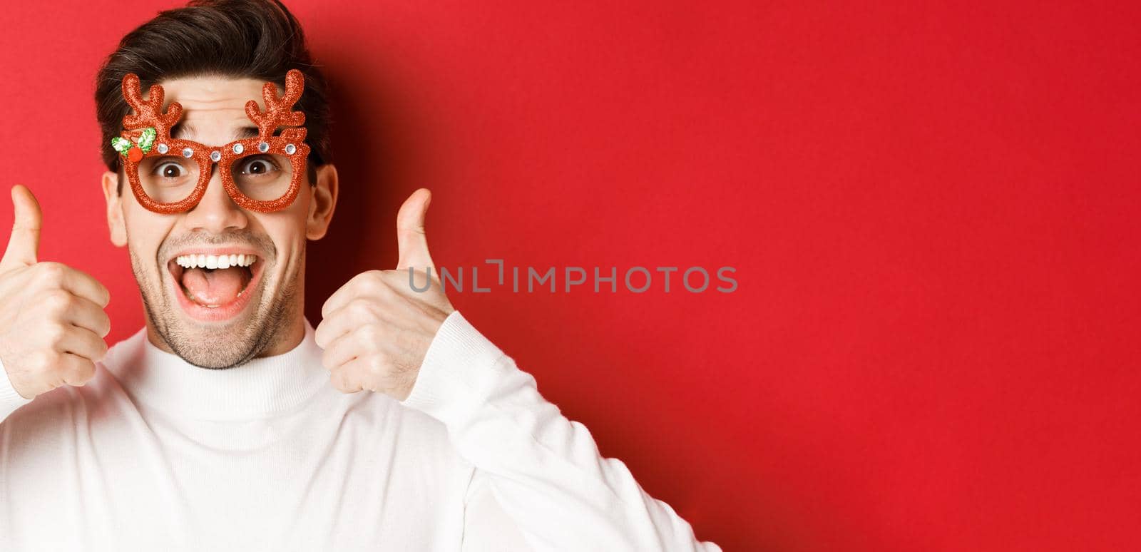 Concept of winter holidays, christmas and celebration. Close-up of excited handsome man in party glasses, smiling and showing thumbs-up in approval, standing over red background.