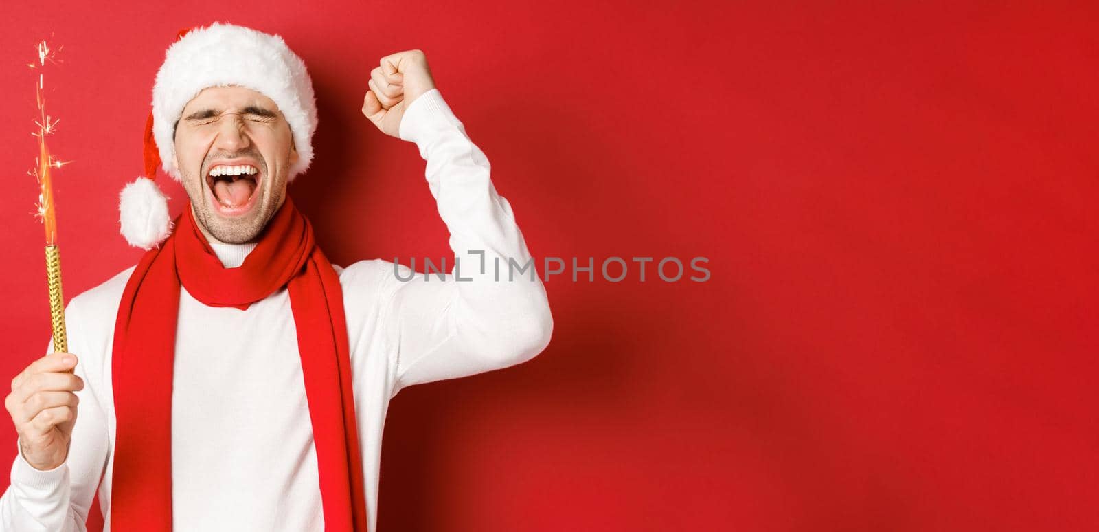 Concept of christmas, winter holidays and celebration. Handsome man celebrating new year and having fun, holding sparkler and smiling, wearing santa hat, standing over red background.