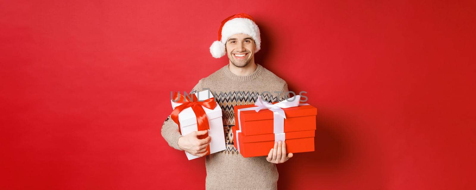 Concept of winter holidays, new year and celebration. Portrait of handsome man in santa hat and sweater, holding boxes with christmas presents and smiling, prepared gifts, red background.