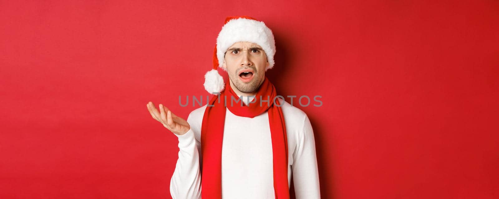 Concept of christmas, winter holidays and celebration. Portrait of confused man in santa hat and scarf, frowning and looking perplexed, standing over red background by Benzoix