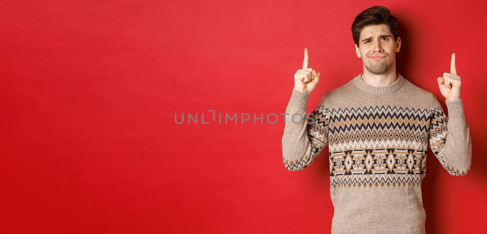 Image of skeptical and unamused handsome guy, showing bad christmas offer, pointing fingers up and grimacing displeased, express dislike, standing over red background.