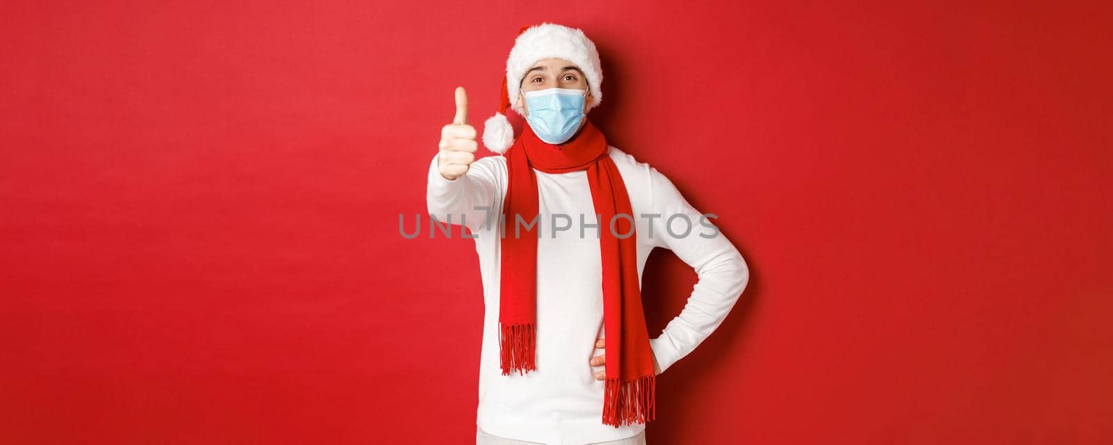 Concept of covid-19, christmas and holidays during pandemic. Cheerful handsome man in medical mask and santa hat, showing thumb-up, celebrating new year and social distancing.