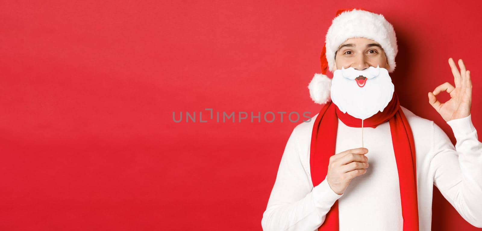 Concept of christmas, winter holidays and celebration. Pleased handsome man in santa hat, holding long white beard mask and showing okay sign, standing over red background.