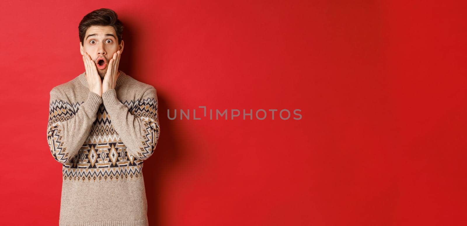 Image of surprised handsome guy reacting to cool new year promo offer, gasping amazed, wearing christmas sweater, standing over red background by Benzoix