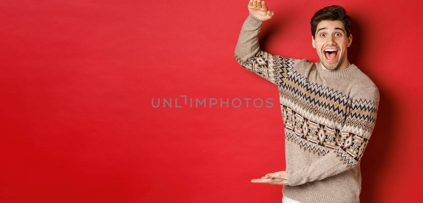 Portrait of excited and happy handsome man in christmas sweater, showing something big, holding large gift for holidays, standing over red background by Benzoix