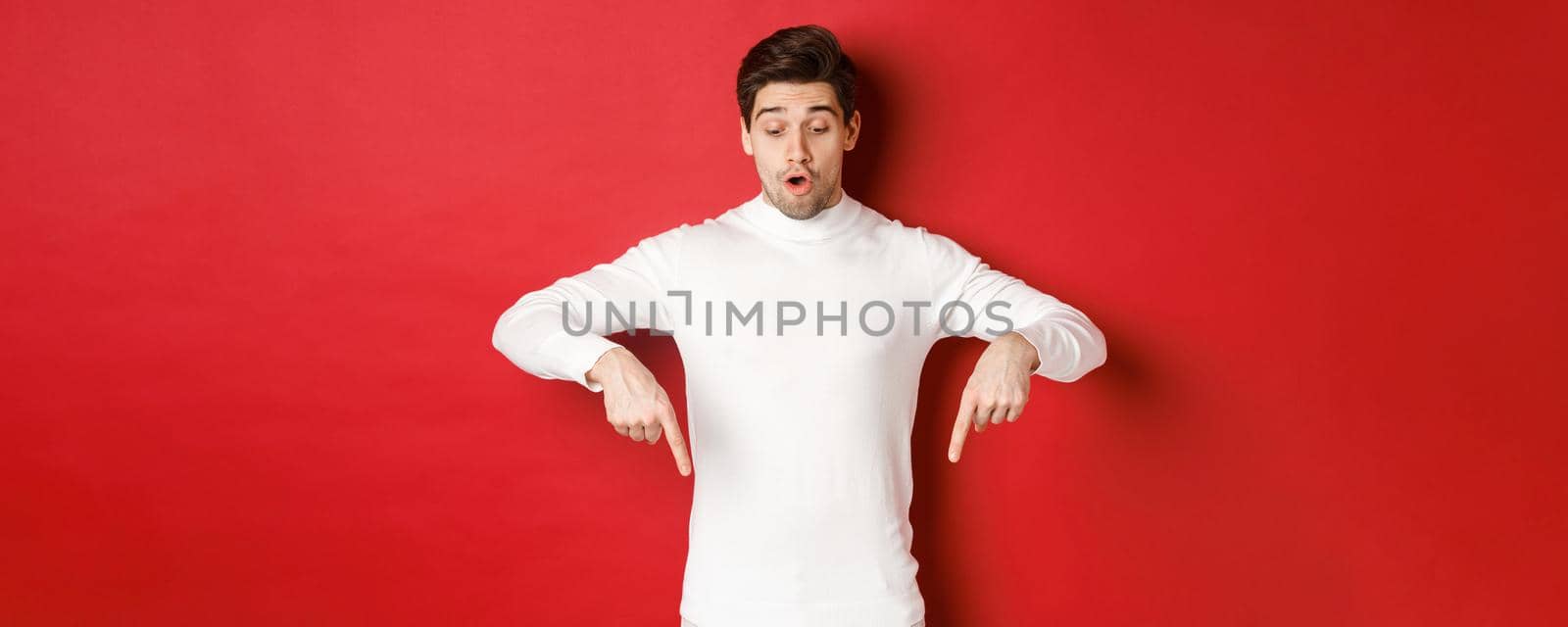 Portrait of surprised good-looking guy in white sweater, looking and pointing fingers down at logo, standing against red background.