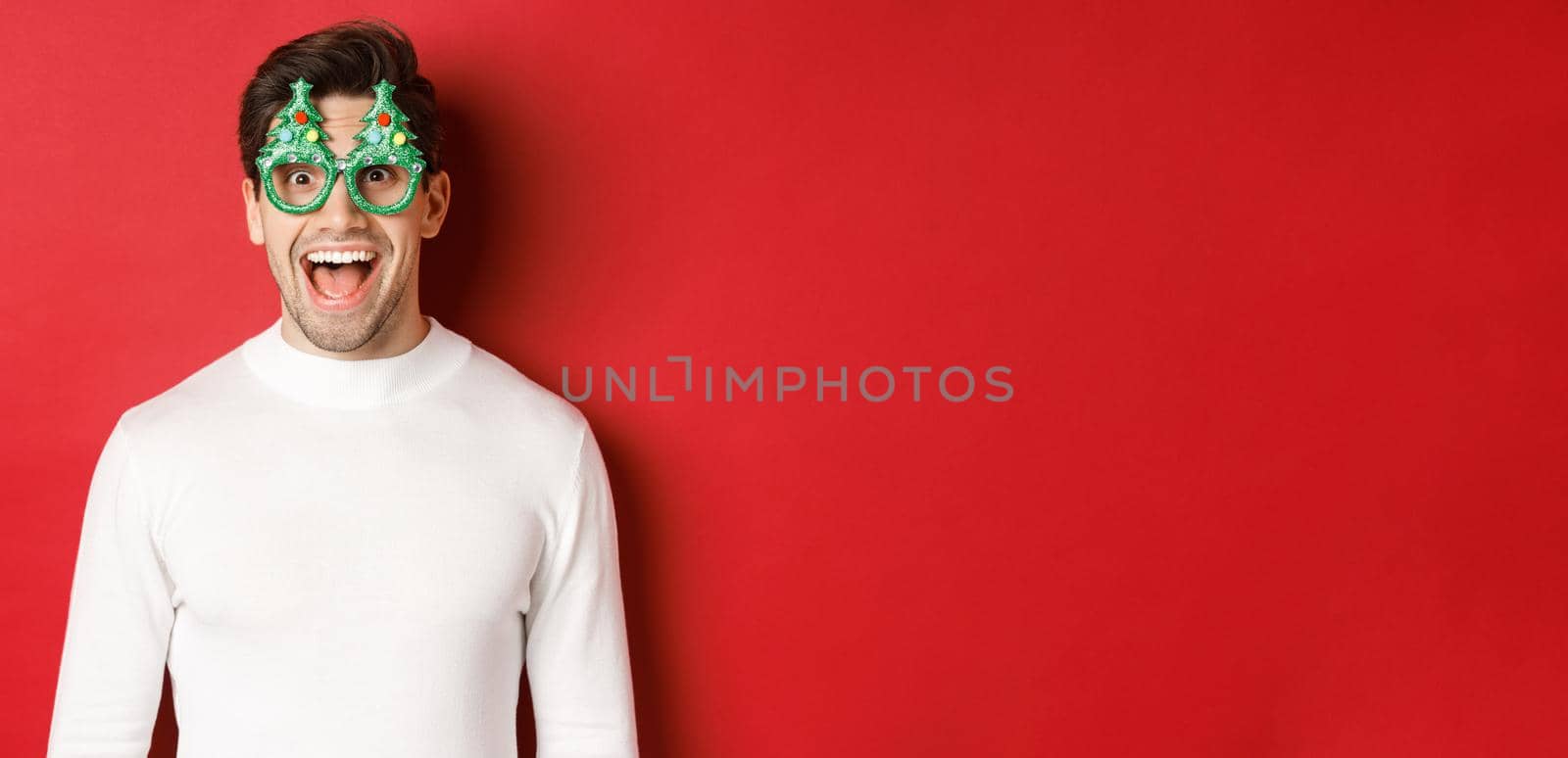 Concept of christmas, winter holidays and celebration. Close-up of handsome young man enjoying new year, wearing party glasses and looking amazed at camera, standing over red background.