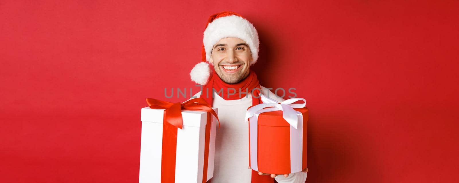 Concept of winter holidays, christmas and lifestyle. Close-up of smiling handsome guy in santa hat and scarf, wishing happy new year and holding gifts, standing with presents over red background by Benzoix