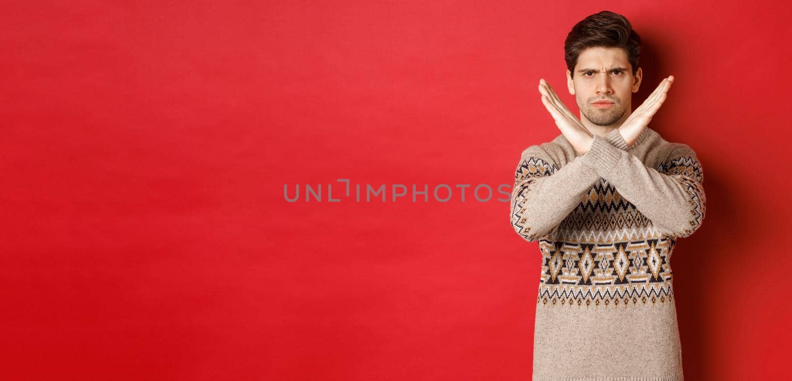 Image of angry and serious handsome man in christmas sweater, telling no or stop, showing cross gesture to restrict you from something, prohibit action, standing over red background.