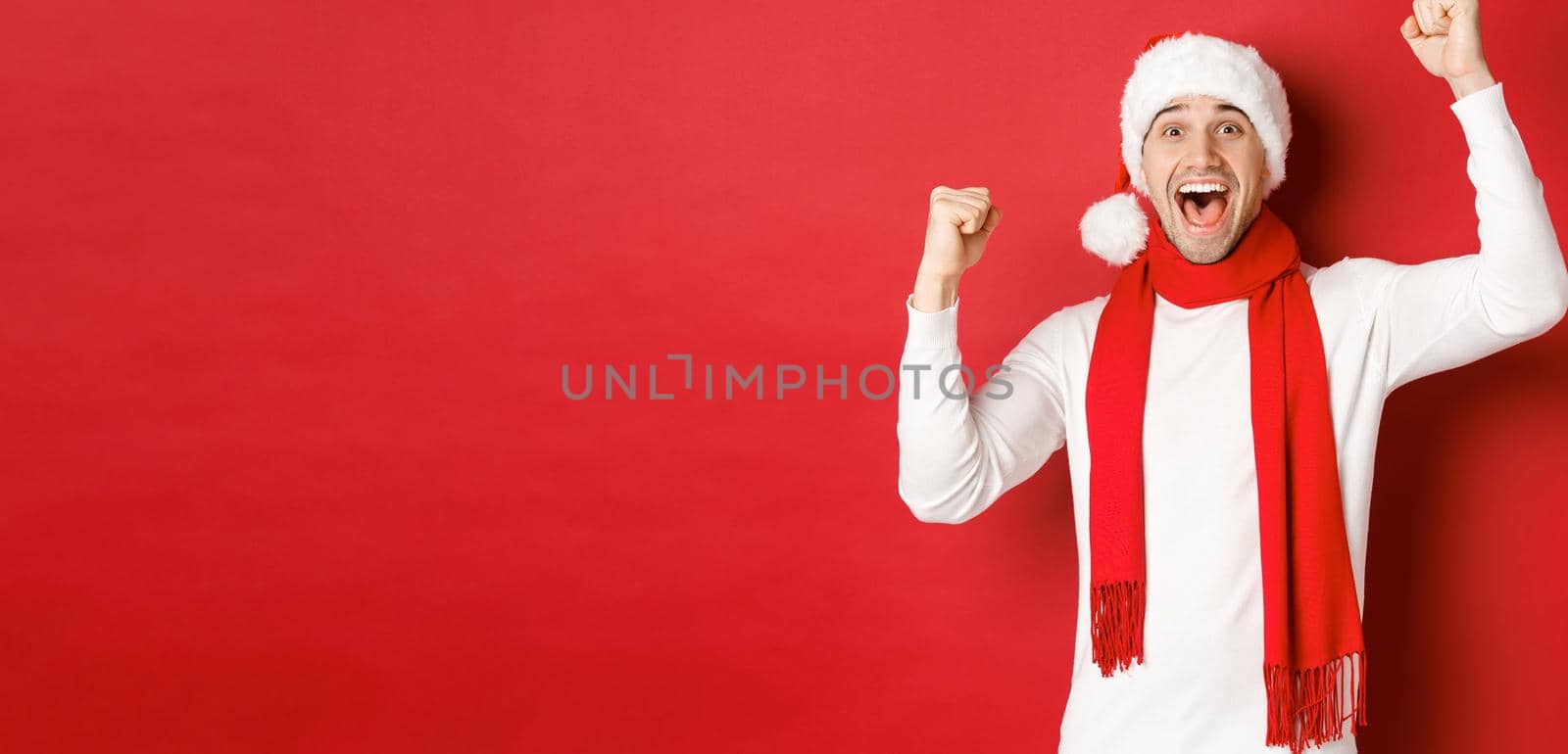 Image of joyful caucasian man in santa hat and scarf, shouting for joy and raising hands, celebrating victory or win, triumphing over red background by Benzoix