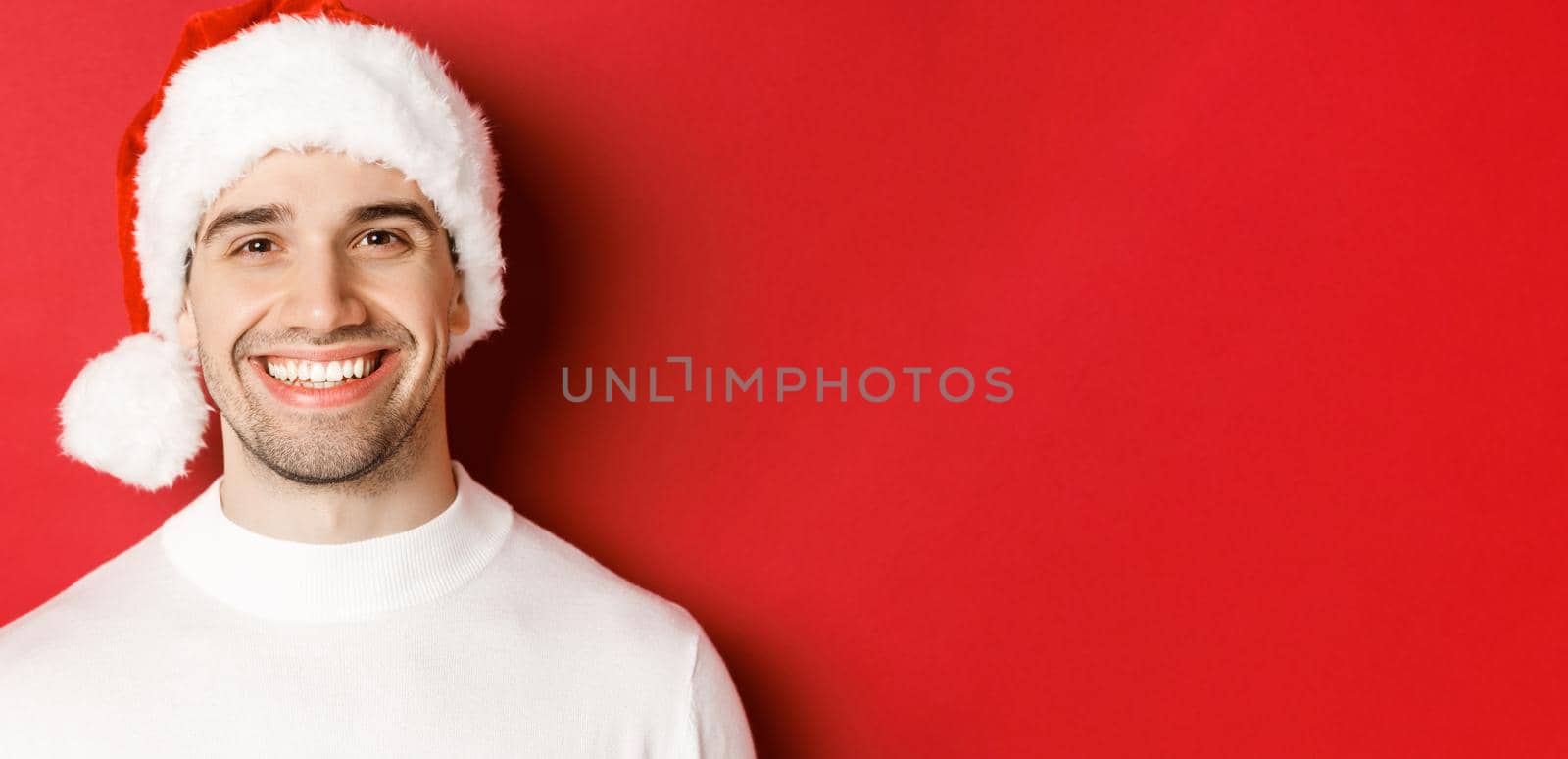 Close-up of attractive smiling man in white sweater and santa hat, looking happy, enjoying winter holidays, standing against red background.
