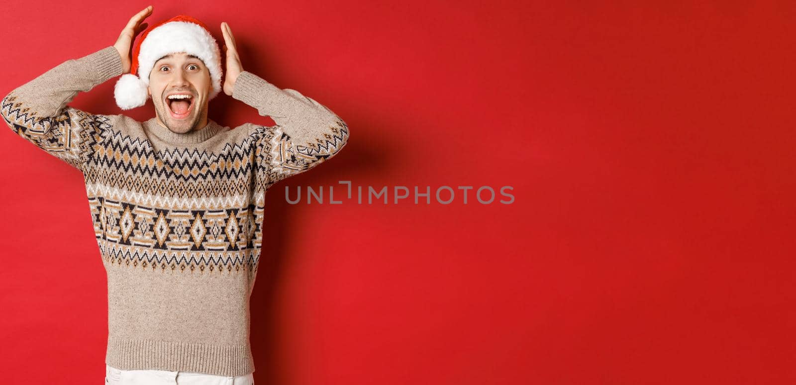 Image of surprised and happy young man in santa hat and christmas sweater, receiving amazing gift, standing excited against red background.