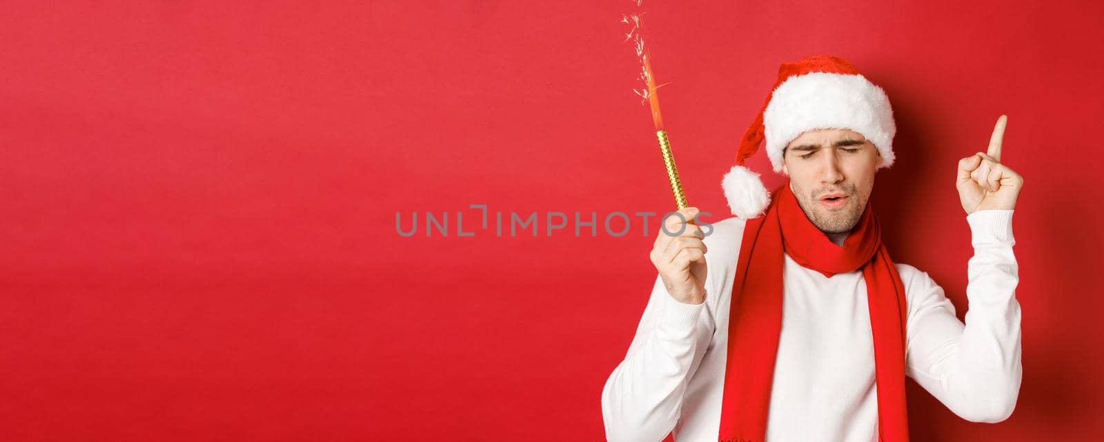 Concept of christmas, winter holidays and celebration. Attractive guy enjoying new year party, dancing with sparkler, wearing santa hat and scarf, standing over red background.