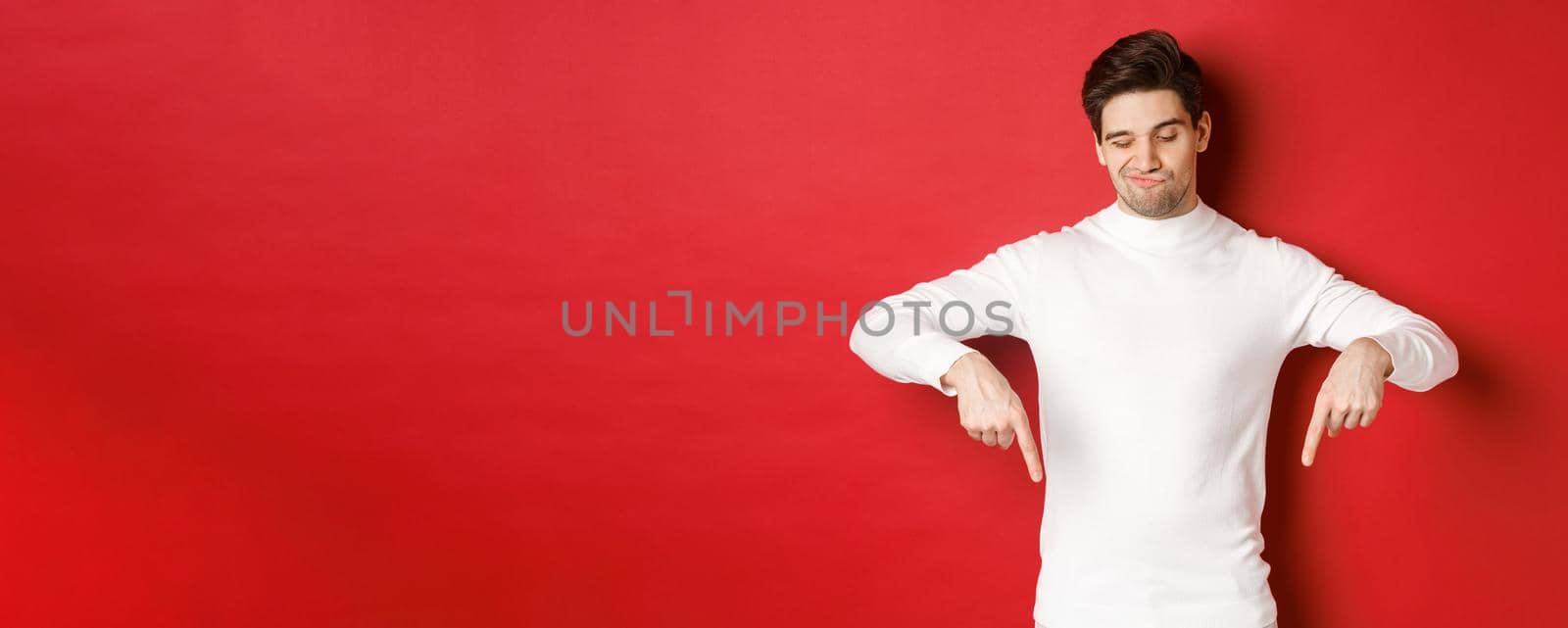 Portrait of unamused handsome man, looking skeptical while pointing fingers down at something bad, standing over red background by Benzoix