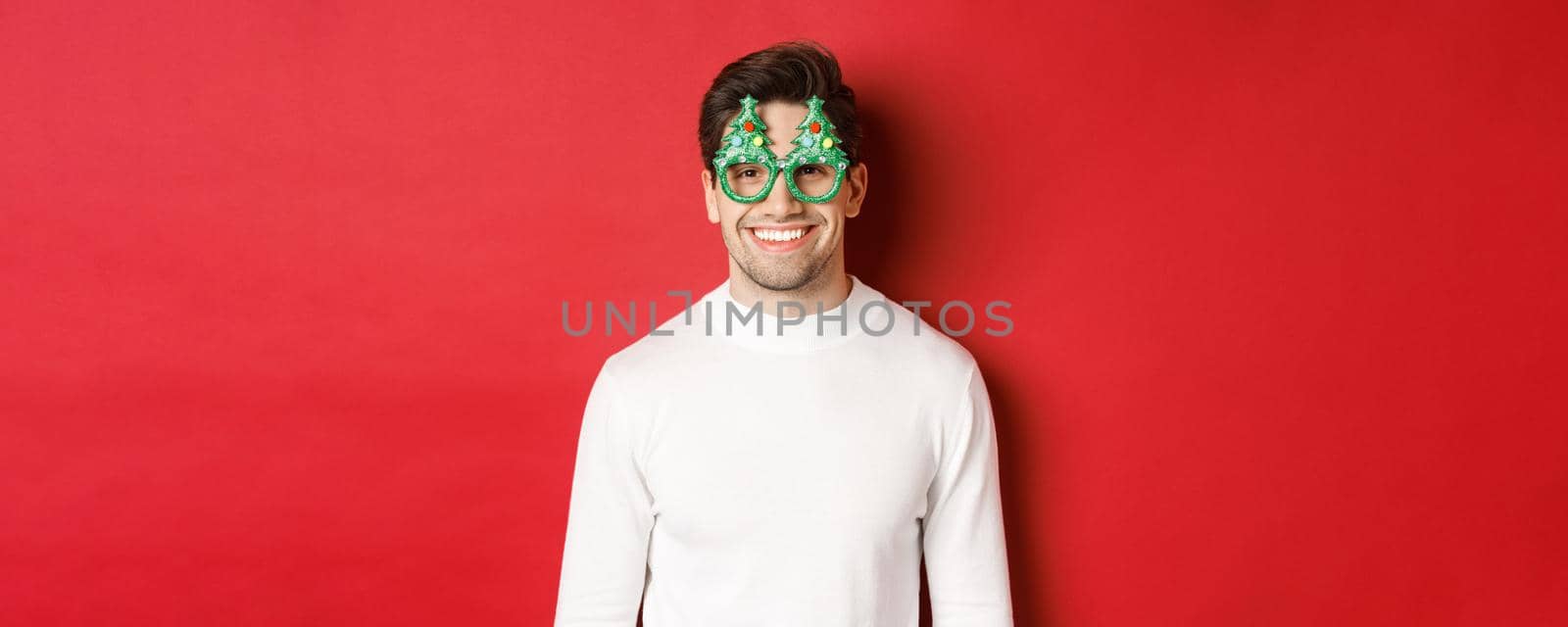 Concept of christmas, winter holidays and celebration. Close-up of handsome man in white sweater and party glasses, enjoying new year, standing over red background.