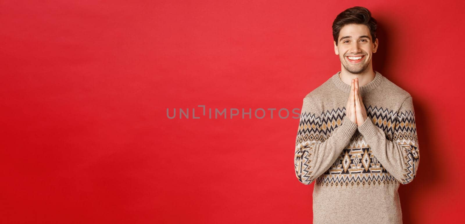 Image of happy and cute man in christmas sweater, asking for favour, holding hands together, smiling and saying thank you, feeling grateful, standing over red background.