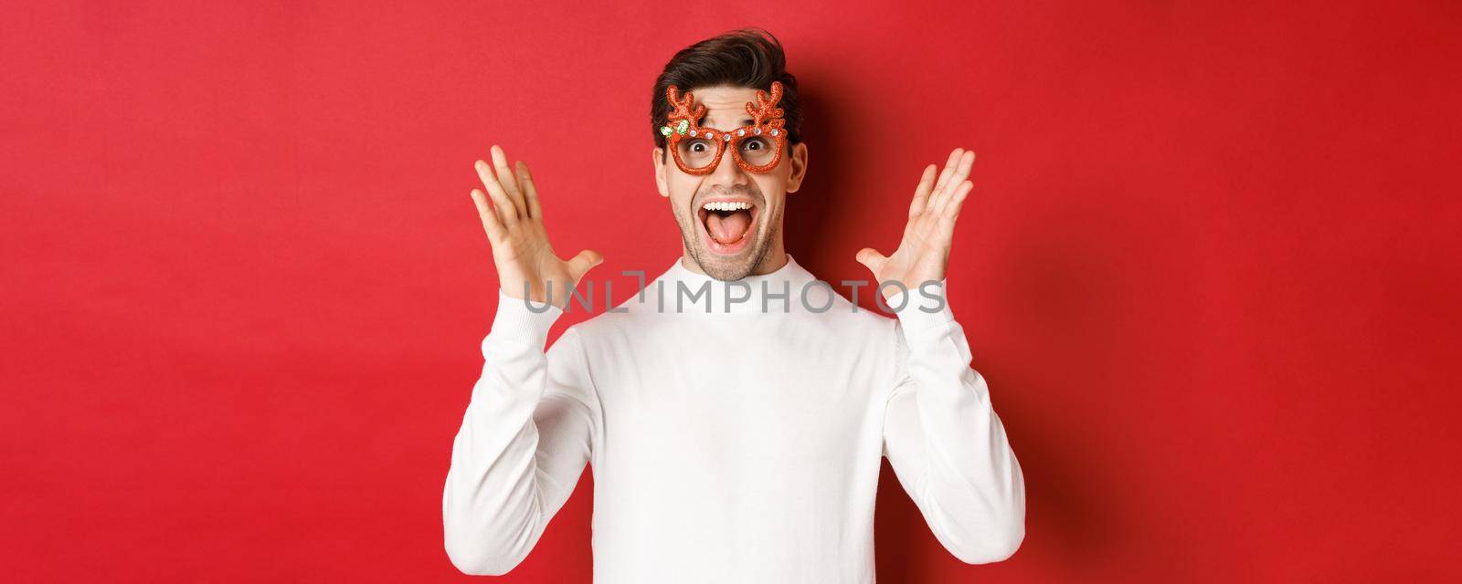 Close-up of surprised handsome guy in white sweater and party glasses, hear amazing christmas promo offer, rejoicing and standing over red background by Benzoix