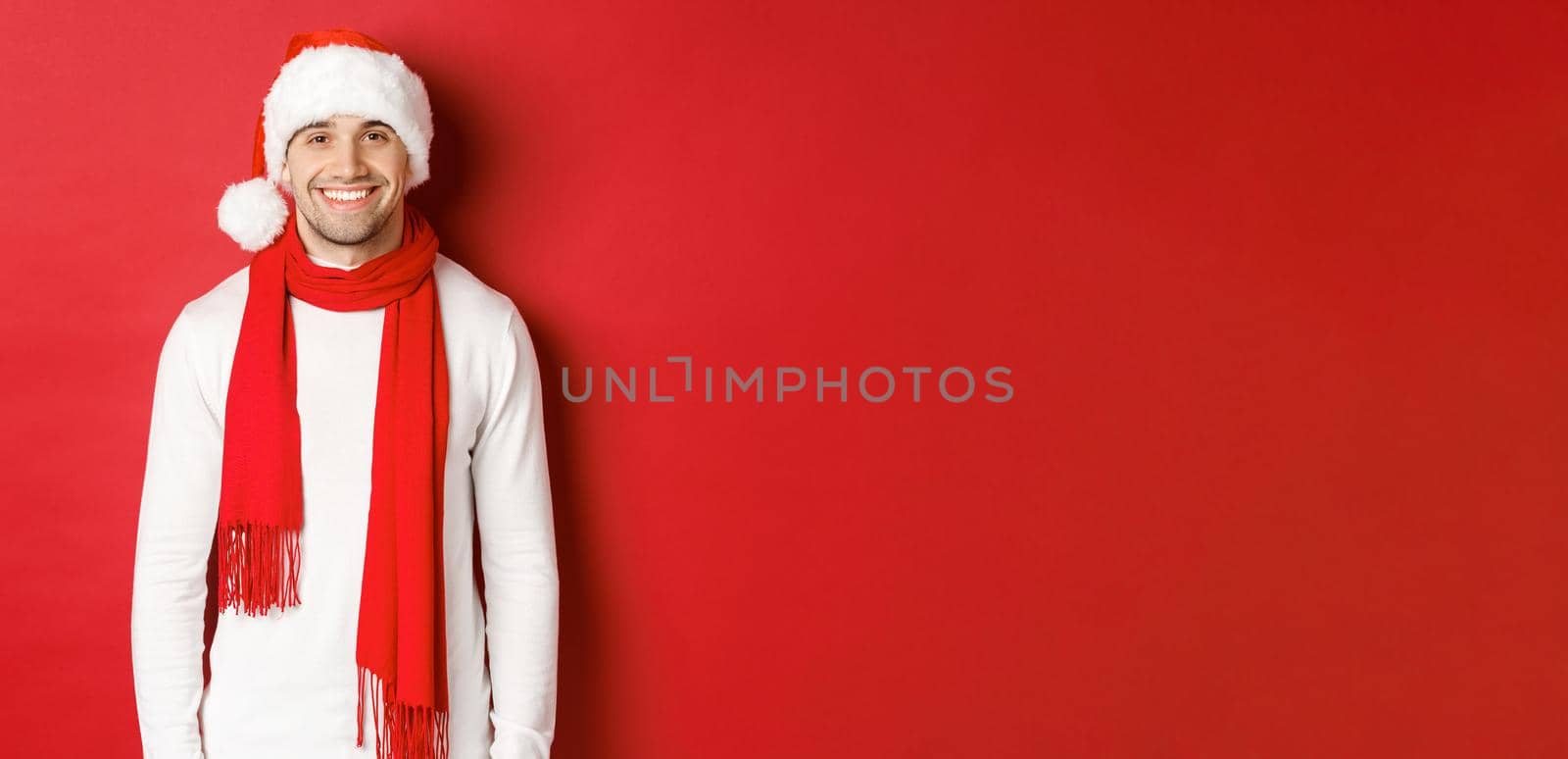 Happy caucasian guy celebrating christmas and smiling, wearing santa hat, scarf and white sweater, enjoying new year holidays, standing over red background by Benzoix