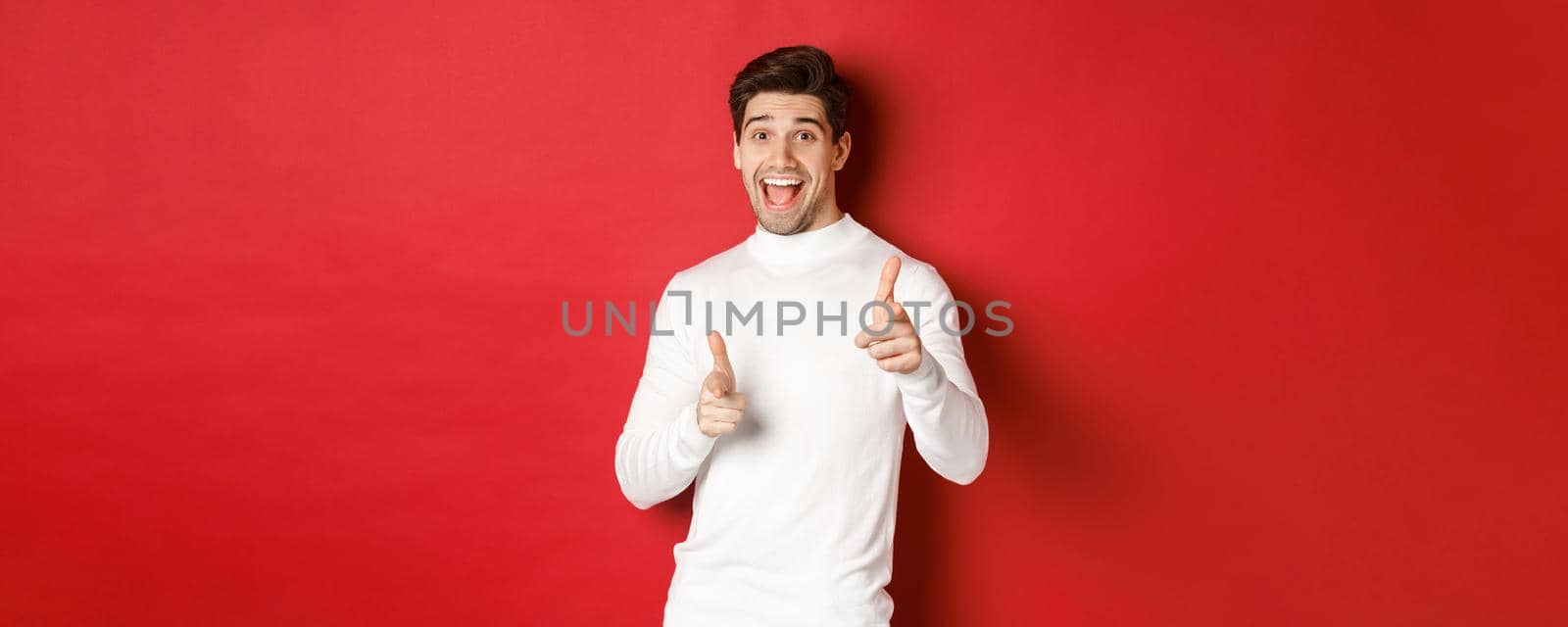 Concept of winter holidays, christmas and lifestyle. Cheerful handsome guy in white sweater congratulating you, pointing fingers at camera and wishing happy new year, standing over red background.