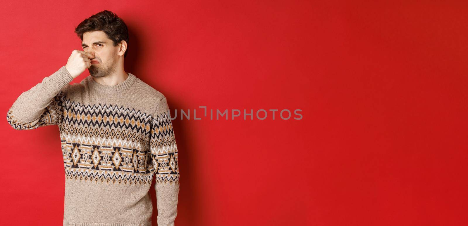 Image of disgusted young man in winter sweater, shut nose and grimacing from bad smell, standing over red background by Benzoix