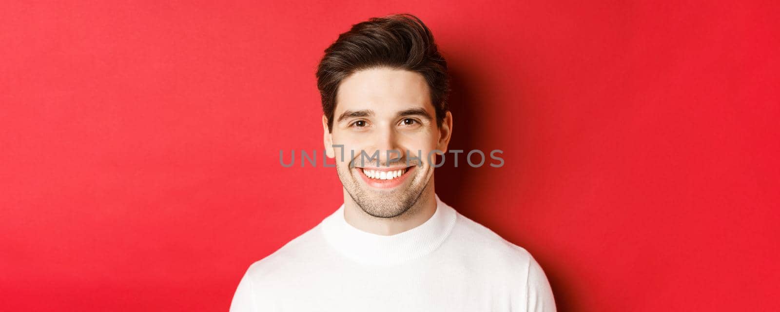 Close-up of handsome smiling brunette man, wearing white sweater, smiling happy and confident, standing against red background.