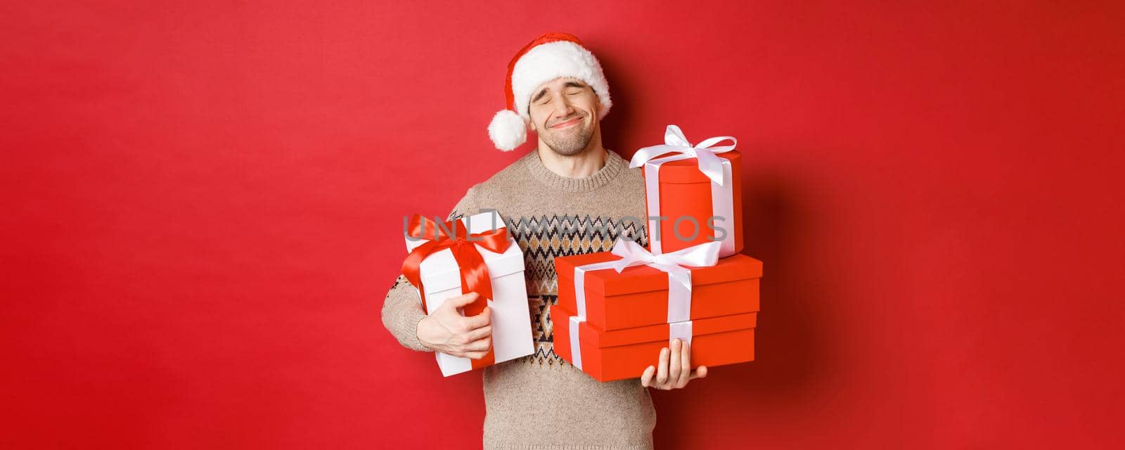 Concept of winter holidays, new year and celebration. Portrait of lovely smiling man receiving pile of presents, holding gifts and being touched with surprise, standing over red background grateful.