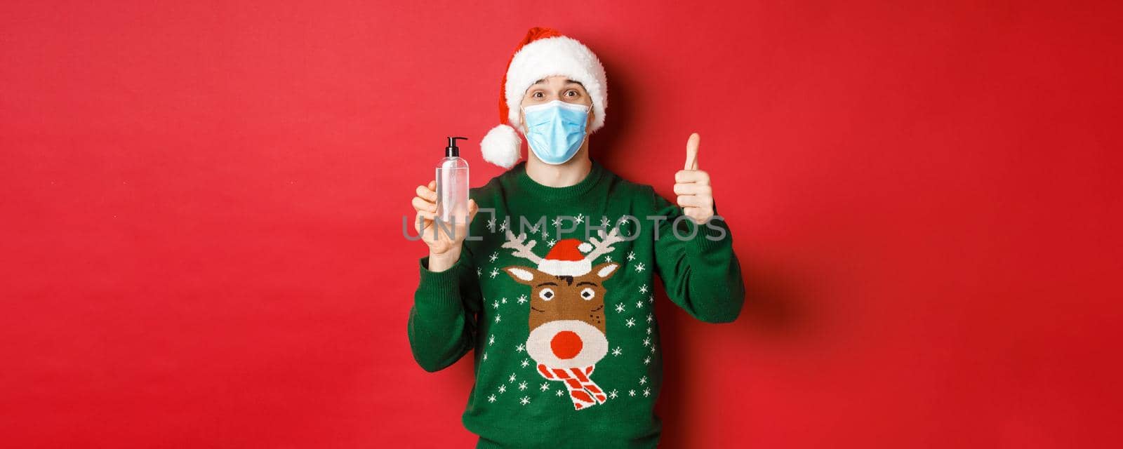 Concept of new year, coronavirus and social distancing. Portrait of cheerful man in santa hat, christmas sweater and medical mask, showing thumb-up and recommending hand sanitizer by Benzoix