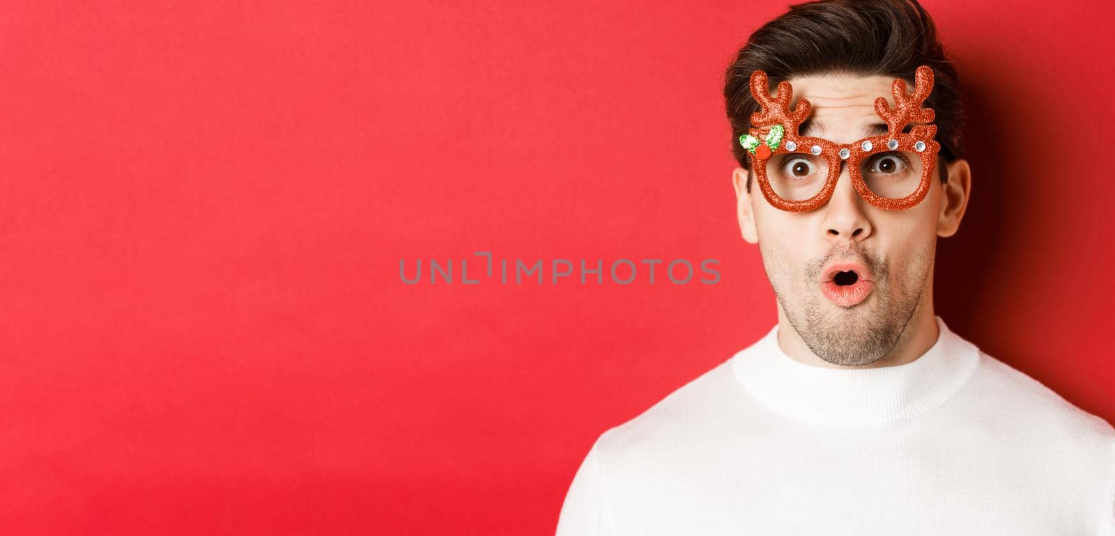 Concept of winter holidays, christmas and celebration. Close-up of attractive guy with bristle, wearing new year party glasses and looking impressed at promo offer, standing over red background by Benzoix