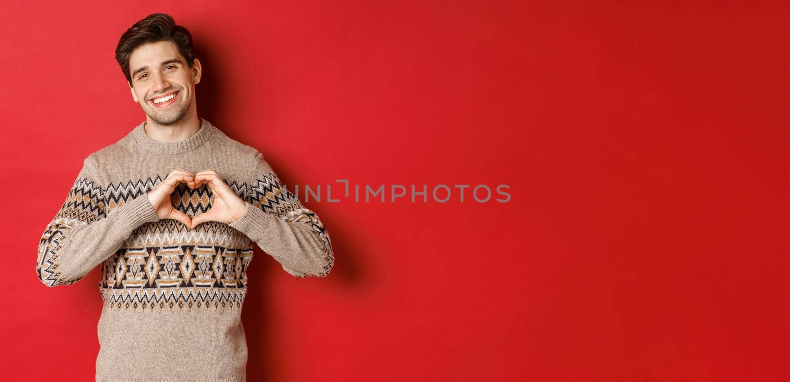 Image of handsome bearded guy in winter sweater, wishing merry christmas and showing heart sign, smiling lovely at camera, standing against red background.