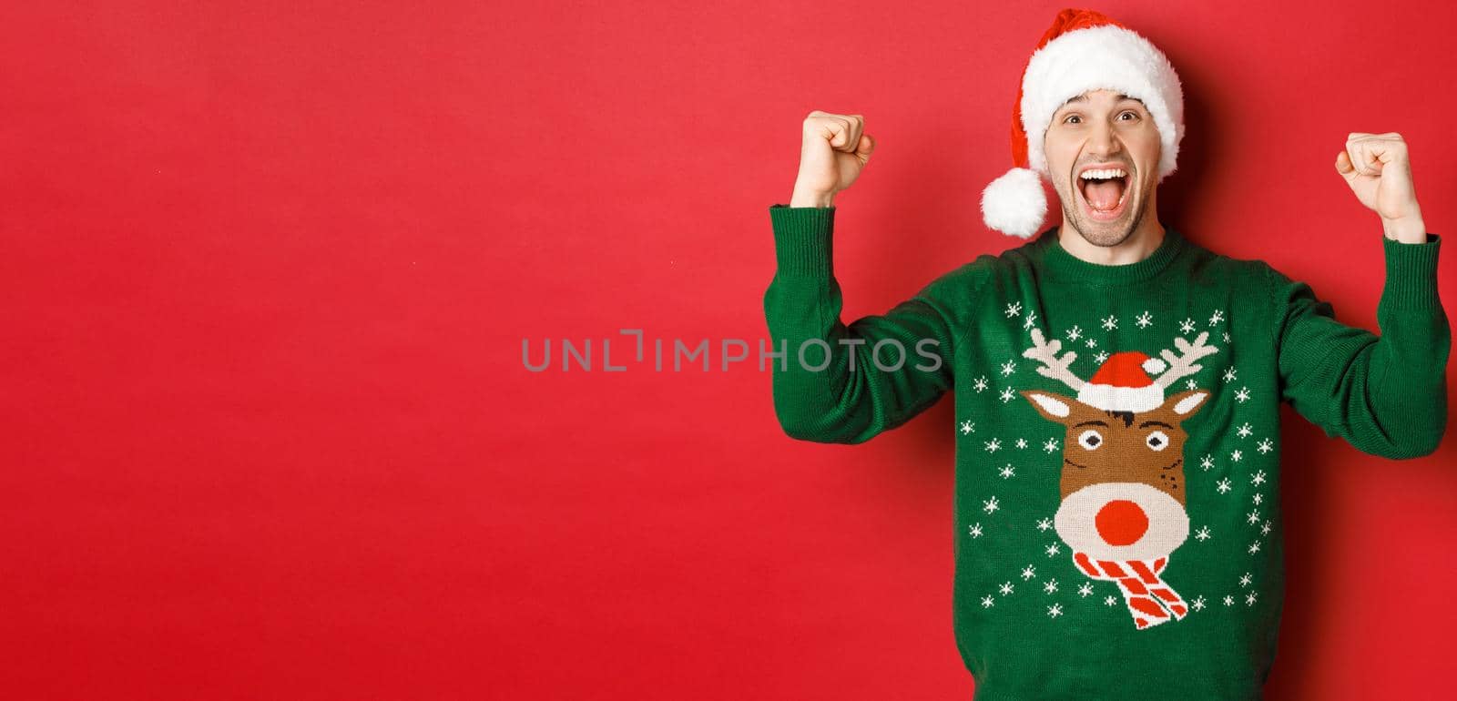 Portrait of cheerful attractive man celebrating new year, wearing green sweater and santa hat, shouting for joy, winning or triumphing, standing over red background.