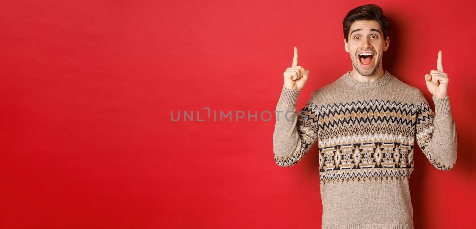 Image of handsome excited man, feeling happy about christmas holidays, pointing fingers up and looking cheerful, showing advertisement, standing in sweater over red background by Benzoix