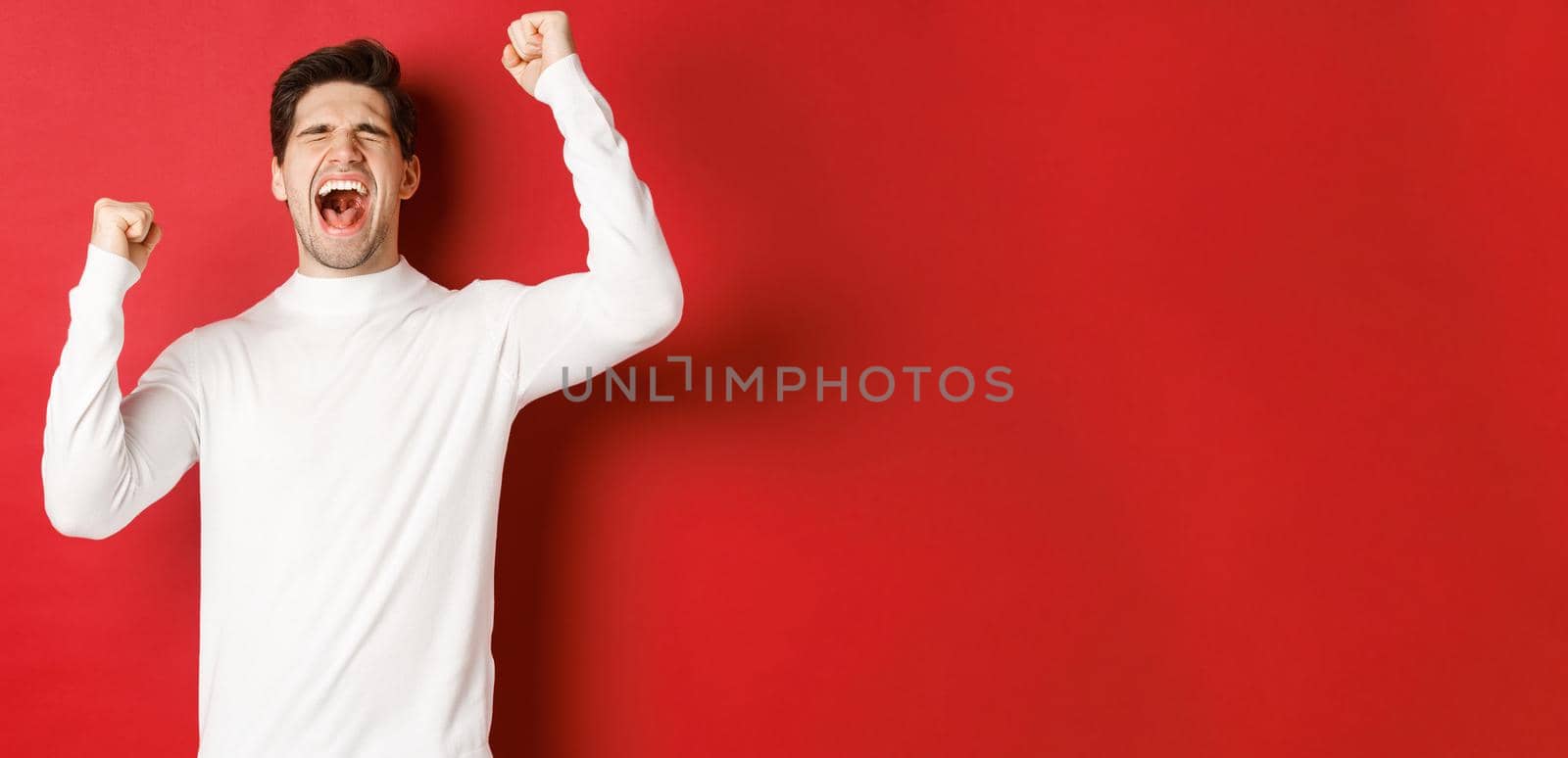 Portrait of handsome man in white sweater, feeling cheerful, celebrating victory, shouting for joy and raising hands up in victory, standing over red background.
