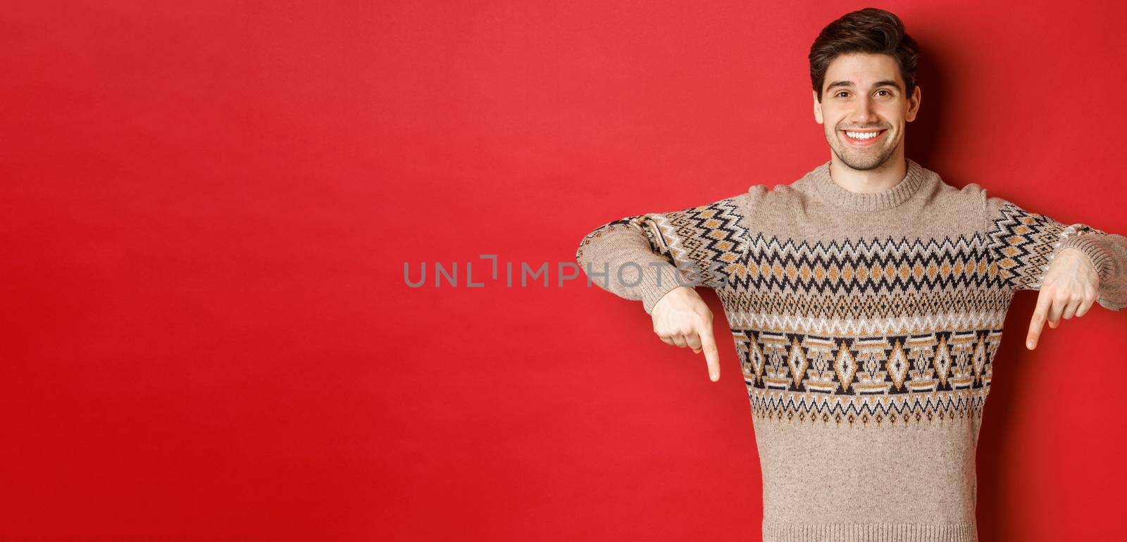 Concept of christmas celebration, winter holidays and lifestyle. Attractive happy man in xmas sweater showing promotion, pointing fingers down at logo, standing over red background by Benzoix